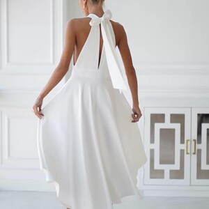 May include: A white halter dress with a flowing skirt and a large bow detail at the back. The dress has a sleeveless design and a midi length. The model is wearing white heeled mules. The background is a white room with a cabinet.