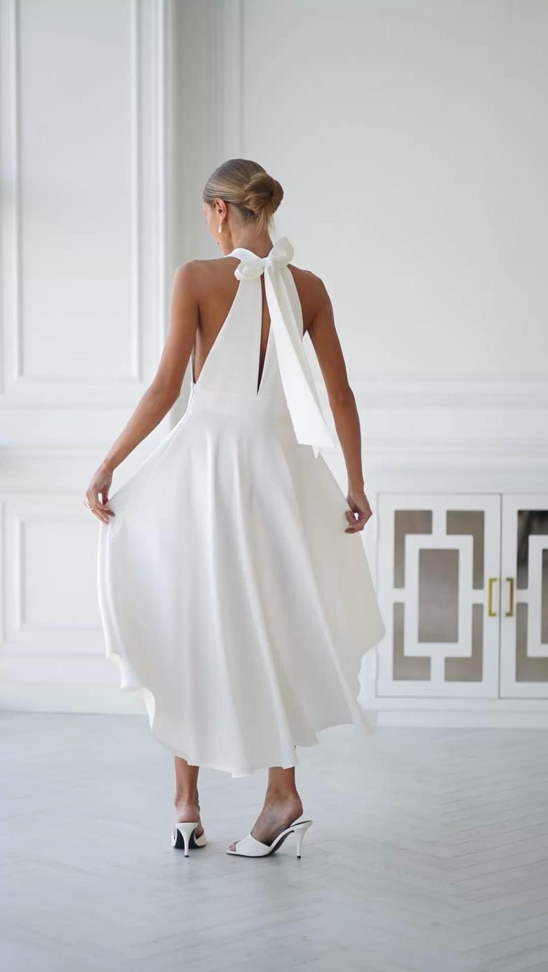 May include: A white halter dress with a flowing skirt and a large bow detail at the back. The dress has a sleeveless design and a midi length. The model is wearing white heeled mules. The background is a white room with a cabinet.