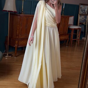 May include: A pale yellow, one-shoulder gown with a flowing cape detail. The dress has a fitted bodice and a floor-length skirt. The fabric appears to be lightweight and flowing. The dress is suitable for formal events.