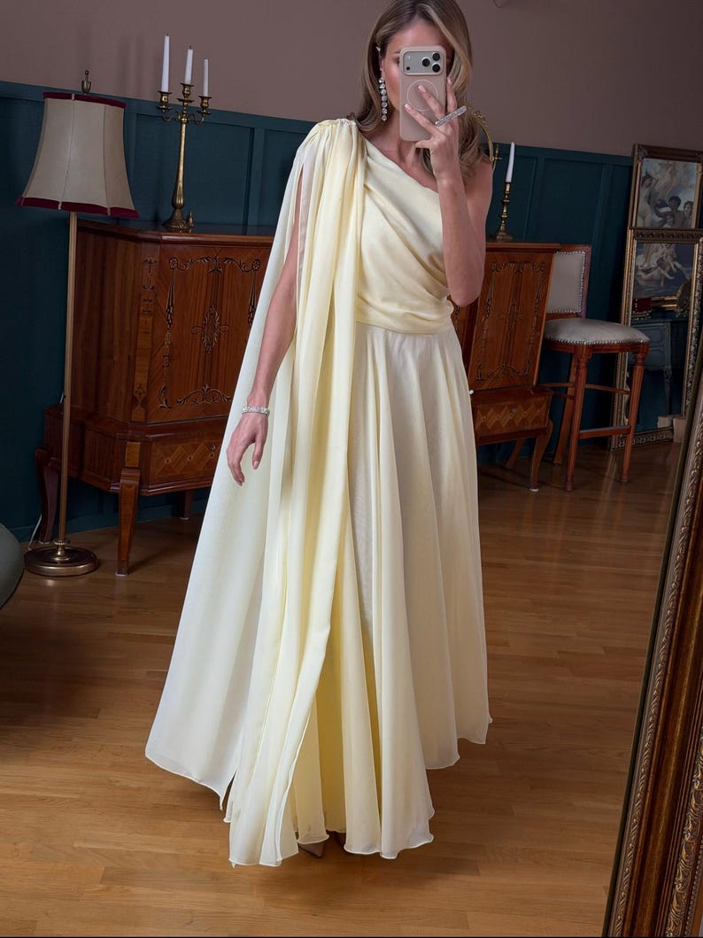 May include: A pale yellow, one-shoulder gown with a flowing cape detail. The dress has a fitted bodice and a floor-length skirt. The fabric appears to be lightweight and flowing. The dress is suitable for formal events.