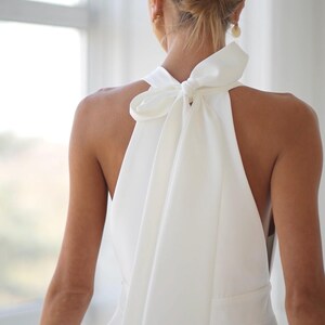 Pu&ograve; includere: Abito bianco con collo all'americana e grande fiocco sulla schiena. L'abito ha un corpetto aderente e una gonna fluida. I capelli della modella sono raccolti in uno chignon basso e indossa orecchini d'oro. L'abito &egrave; realizzato in tessuto liscio, forse satin.