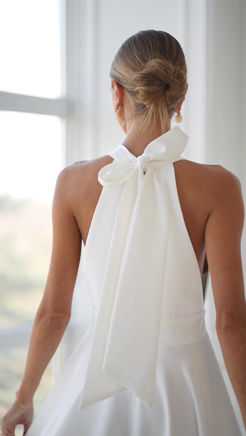 Pu&ograve; includere: Abito bianco con collo all'americana e grande fiocco sulla schiena. L'abito ha un corpetto aderente e una gonna fluida. I capelli della modella sono raccolti in uno chignon basso e indossa orecchini d'oro. L'abito &egrave; realizzato in tessuto liscio, forse satin.