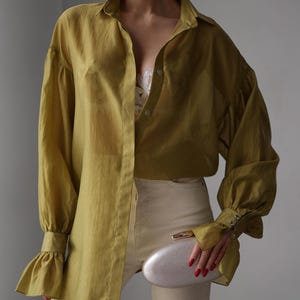 Puede incluir: Camisa verde oliva con mangas largas y abullonadas y pu&ntilde;os con volantes. La camisa se combina con pantalones color crema y un clutch plateado. El conjunto tiene un estilo minimalista.