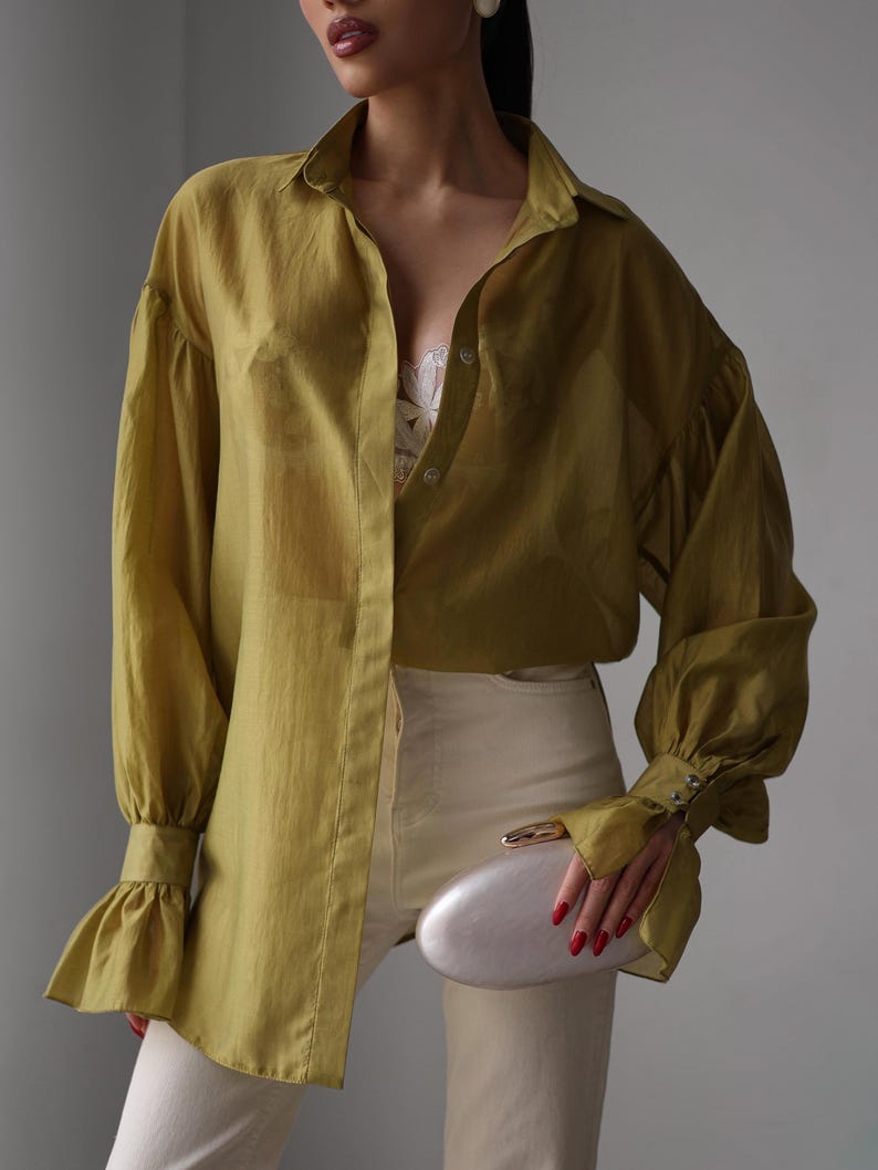 Puede incluir: Camisa verde oliva con mangas largas y abullonadas y pu&ntilde;os con volantes. La camisa se combina con pantalones color crema y un clutch plateado. El conjunto tiene un estilo minimalista.