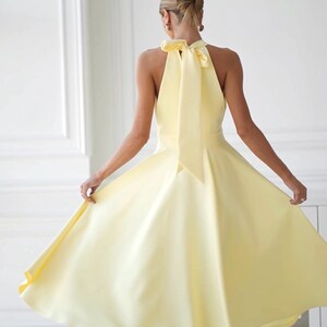May include: A pale yellow halter dress with a flowing skirt. The dress features a large bow at the back of the neck. The model is wearing white heels. The dress is a solid color and appears to be made of a smooth fabric.