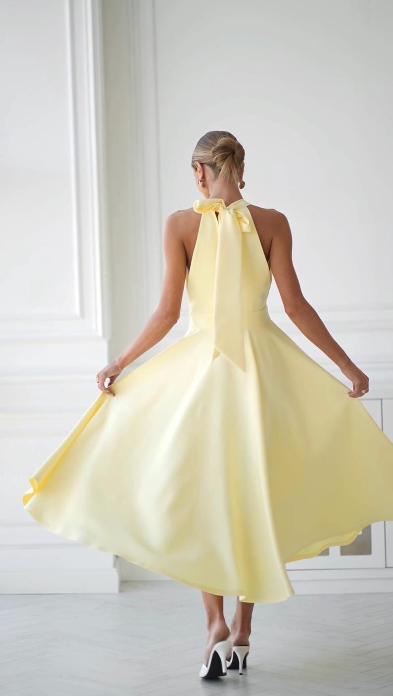 May include: A pale yellow halter dress with a flowing skirt. The dress features a large bow at the back of the neck. The model is wearing white heels. The dress is a solid color and appears to be made of a smooth fabric.