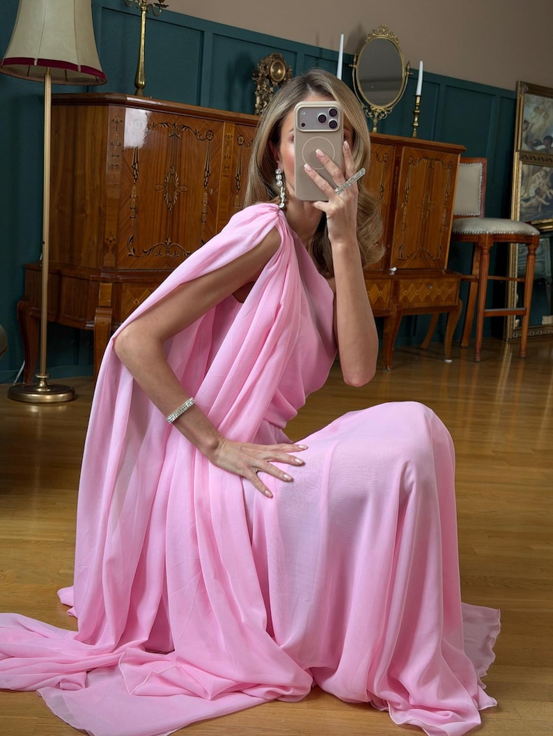 May include: A flowing, light pink dress with a cape-like design. The dress is made of a soft, draping fabric and is floor-length. The model is wearing a bracelet and earrings. The background includes a wooden cabinet, a mirror, and a floor lamp.