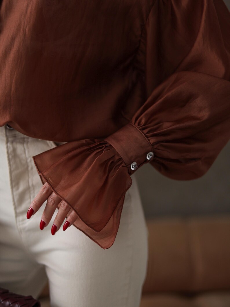 Puede incluir: Primer plano de una blusa marr&oacute;n con mangas largas y fluidas. El pu&ntilde;o de la manga est&aacute; detallado con dos botones plateados. La blusa se combina con pantalones blancos rotos. Las u&ntilde;as est&aacute;n pintadas de rojo.