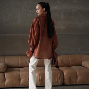 Puede incluir: Una blusa marr&oacute;n con mangas largas y voluminosas y pu&ntilde;os abotonados, combinada con vaqueros blancos con dobladillo deshilachado. El conjunto se completa con tacones burdeos. La modelo lleva pendientes de aro dorados.