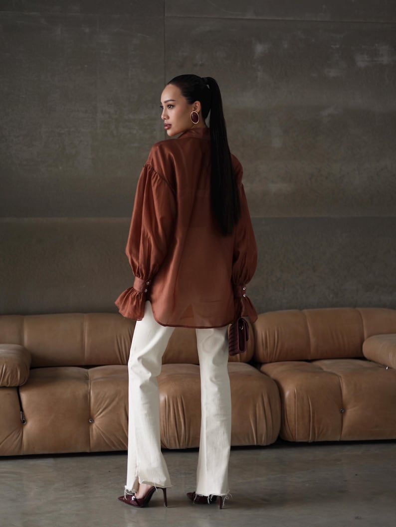 Puede incluir: Una blusa marr&oacute;n con mangas largas y voluminosas y pu&ntilde;os abotonados, combinada con vaqueros blancos con dobladillo deshilachado. El conjunto se completa con tacones burdeos. La modelo lleva pendientes de aro dorados.