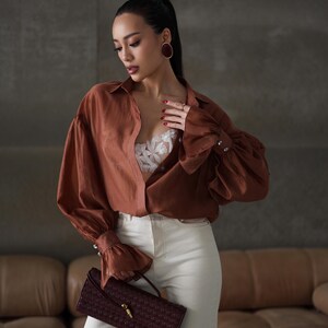 Puede incluir: Una mujer con una camisa abotonada color &oacute;xido con mangas voluminosas y vaqueros blancos. Sostiene un bolso de mano burdeos tejido con un cierre dorado. El conjunto se complementa con pendientes ovalados grandes.