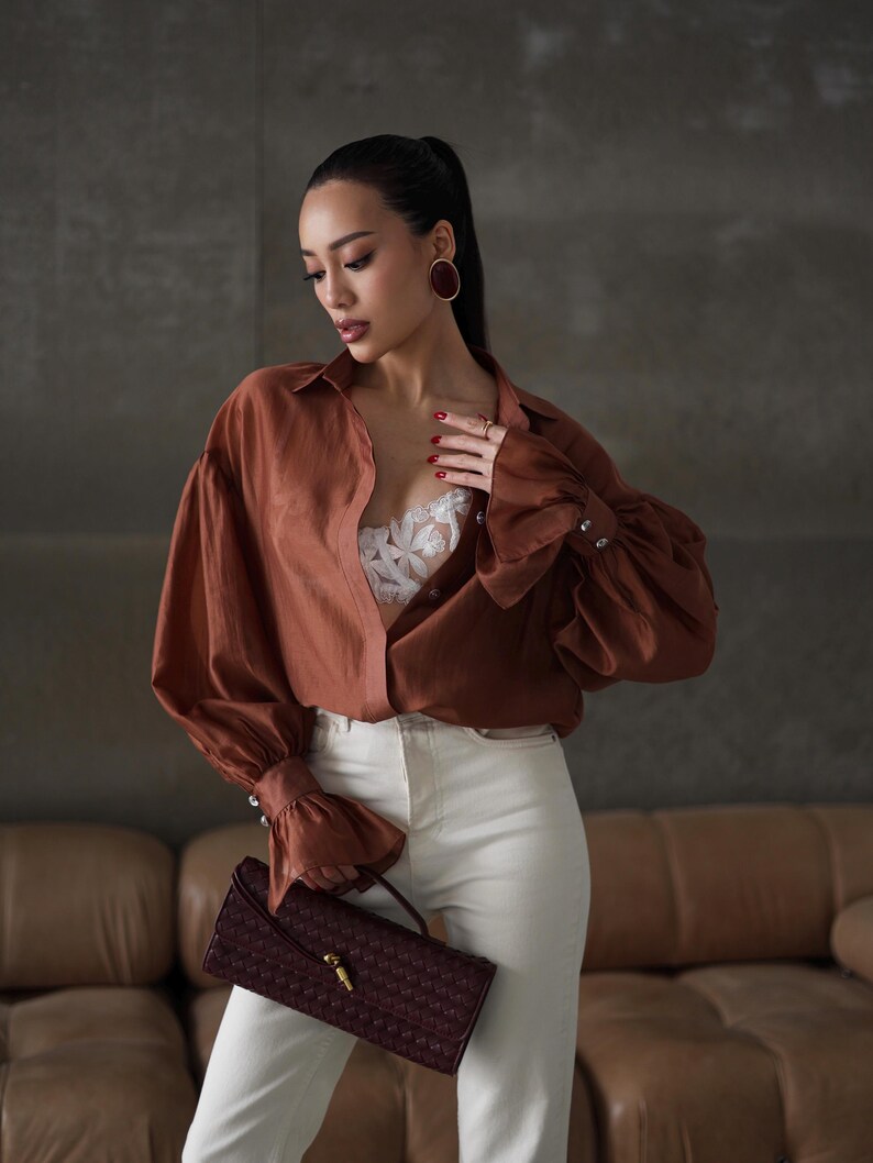 Puede incluir: Una mujer con una camisa abotonada color &oacute;xido con mangas voluminosas y vaqueros blancos. Sostiene un bolso de mano burdeos tejido con un cierre dorado. El conjunto se complementa con pendientes ovalados grandes.