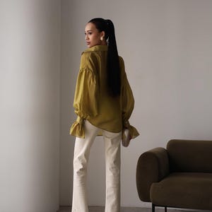 Puede incluir: Blusa verde oliva de manga larga con pu&ntilde;os con volantes y cuello a juego. La blusa se combina con vaqueros acampanados color crema con abertura en el dobladillo. La modelo lleva tacones blancos y un peque&ntilde;o clutch blanco roto.