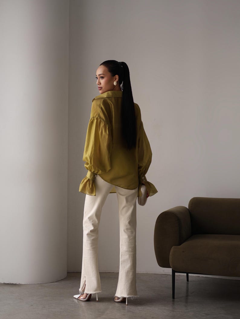 Puede incluir: Blusa verde oliva de manga larga con pu&ntilde;os con volantes y cuello a juego. La blusa se combina con vaqueros acampanados color crema con abertura en el dobladillo. La modelo lleva tacones blancos y un peque&ntilde;o clutch blanco roto.