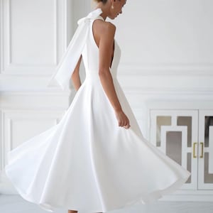 Pu&ograve; includere: Un abito bianco senza maniche con gonna fluida e dettaglio a fiocco sulla schiena. L'abito &egrave; lungo fino al ginocchio e abbinato a tacchi bianchi. L'immagine &egrave; ambientata in uno spazio luminoso e minimalista, che suggerisce un matrimonio o un'occasione speciale.