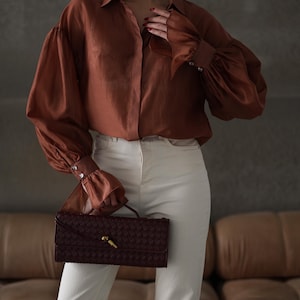 Puede incluir: Una blusa color &oacute;xido con mangas voluminosas y pu&ntilde;os abotonados, combinada con pantalones color crema. Un clutch burdeos tejido con un cierre dorado se sostiene al frente. El conjunto se presenta sobre un fondo neutro.