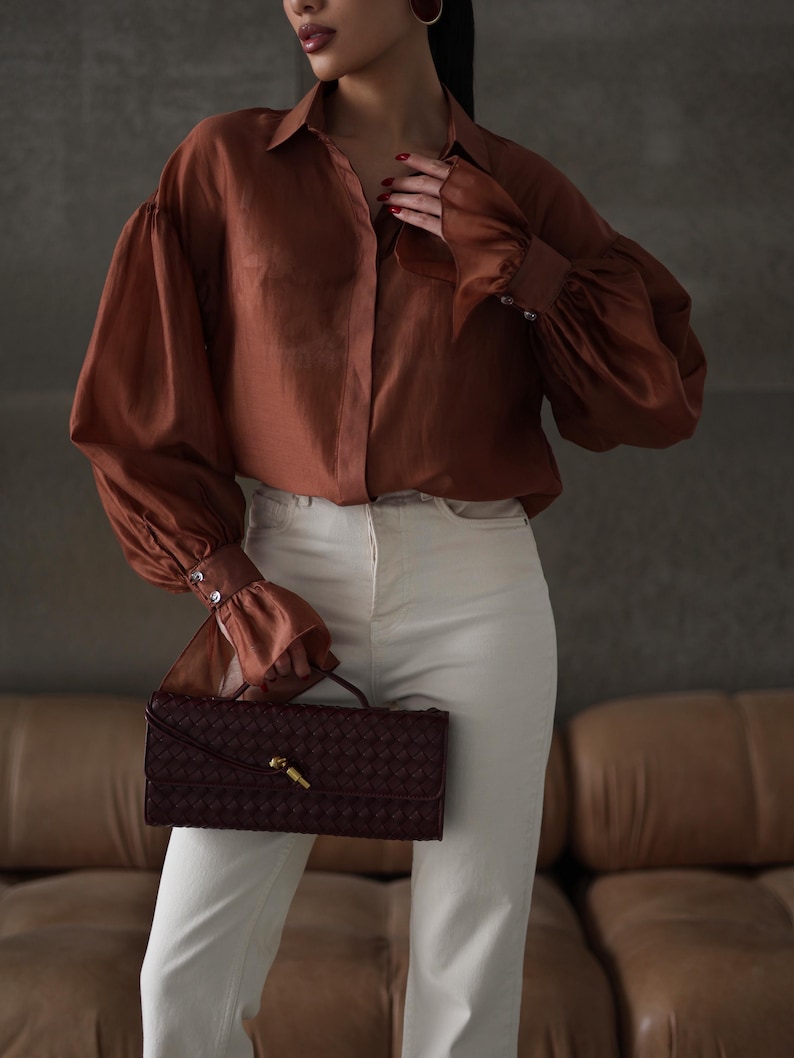 Puede incluir: Una blusa color &oacute;xido con mangas voluminosas y pu&ntilde;os abotonados, combinada con pantalones color crema. Un clutch burdeos tejido con un cierre dorado se sostiene al frente. El conjunto se presenta sobre un fondo neutro.