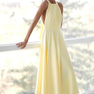 Pu&ograve; includere: Abito giallo pallido con collo all'americana e gonna svasata. L'abito presenta una schiena scoperta e un dettaglio a fiocco. La modella indossa tacchi bianchi. L'abito &egrave; adatto a eventi formali.
