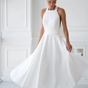 Pu&ograve; includere: Abito midi bianco con collo all'americana e gonna fluida. L'abito presenta uno scollo quadrato e un corpetto aderente. La modella indossa sandali bianchi con tacco alto. L'abito &egrave; adatto per un matrimonio o un'occasione speciale.