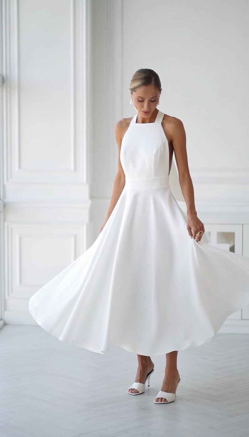 Pu&ograve; includere: Abito midi bianco con collo all'americana e gonna fluida. L'abito presenta uno scollo quadrato e un corpetto aderente. La modella indossa sandali bianchi con tacco alto. L'abito &egrave; adatto per un matrimonio o un'occasione speciale.