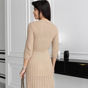 May include: A beige knit dress with a textured bodice and a pleated skirt. The dress has three-quarter sleeves and a fitted waist. A black quilted handbag with a gold chain strap is held in the hand.