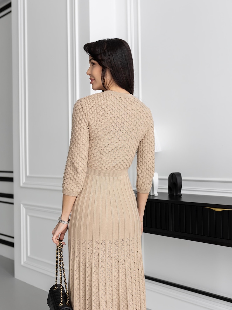 May include: A beige knit dress with a textured bodice and a pleated skirt. The dress has three-quarter sleeves and a fitted waist. A black quilted handbag with a gold chain strap is held in the hand.
