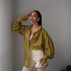 Puede incluir: Una camisa verde oliva con mangas largas y voluminosas, combinada con pantalones blancos. El conjunto se complementa con un clutch plateado. La persona lleva pendientes blancos y tiene el pelo recogido.