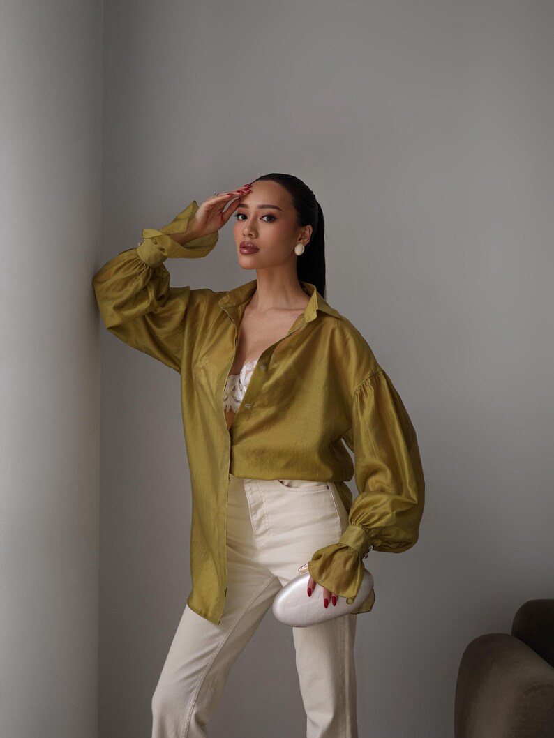 Puede incluir: Una camisa verde oliva con mangas largas y voluminosas, combinada con pantalones blancos. El conjunto se complementa con un clutch plateado. La persona lleva pendientes blancos y tiene el pelo recogido.