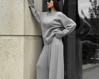 May include: A light gray knitwear set consisting of a turtleneck sweater with a pocket and wide-leg pleated pants. The outfit is paired with a black beret and sunglasses. The ensemble is a monochromatic, modern style.