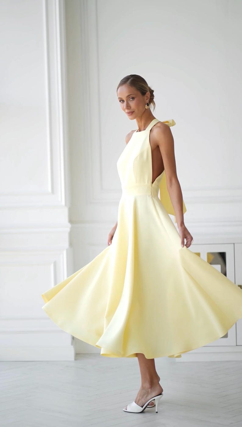 May include: A light yellow halter dress with a flowing skirt. The dress features a bow detail at the back and is knee-length. The model is wearing white heels.