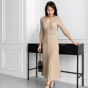 May include: A beige knitted dress with a button-down front, three-quarter sleeves, and a midi-length skirt. The dress features textured knit detailing and small pockets on the chest. The person is wearing black high heels and carrying a black purse.