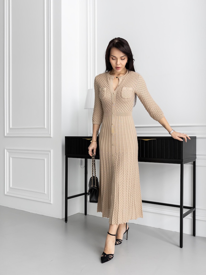 May include: A beige knitted dress with a button-down front, three-quarter sleeves, and a midi-length skirt. The dress features textured knit detailing and small pockets on the chest. The person is wearing black high heels and carrying a black purse.