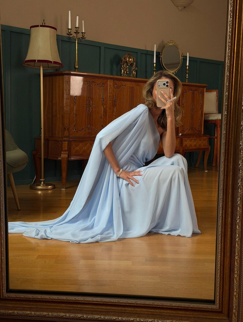 May include: A floor-length, light blue, one-shoulder gown featuring a draped bodice and a flowing, floor-sweeping skirt. The garment is crafted from a lightweight, semi-sheer fabric that creates soft folds and a cascading silhouette.