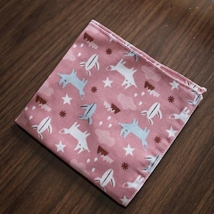 May include: A square fabric cloth in a dusty pink hue featuring a repeating pattern of stylized white and light blue foxes, small white stars, brown mountain shapes, and tiny brown flower-like icons, finished with a stitched hem along the edges.