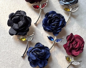 May include: A collection of six flower brooches in various colors, including burgundy, navy, black, and blue-grey. Each brooch features a fabric flower with a silver-tone stem and leaf accents adorned with rhinestones and colored gems.