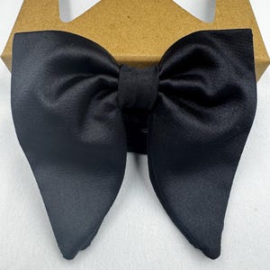 May include: A black satin bow tie in a butterfly shape. The bow tie is neatly tied and rests on a brown cardboard box, creating a contrast in textures and colours. The bow tie is a formal accessory.