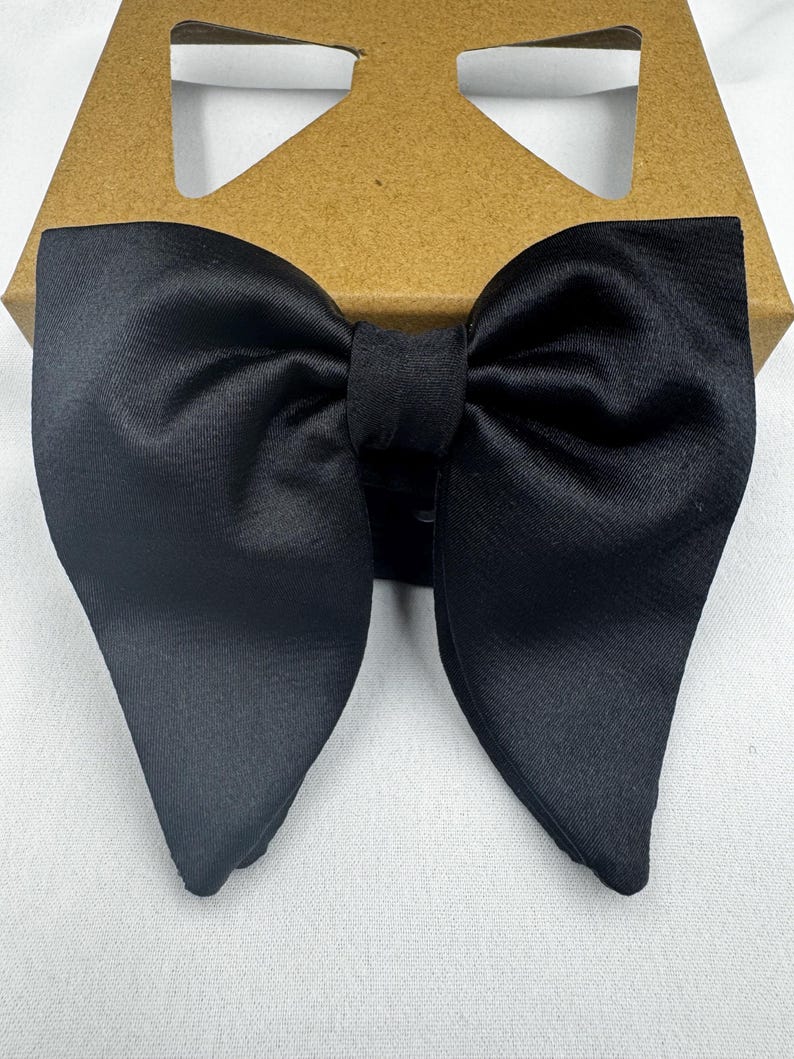 May include: A black satin bow tie in a butterfly shape. The bow tie is neatly tied and rests on a brown cardboard box, creating a contrast in textures and colours. The bow tie is a formal accessory.