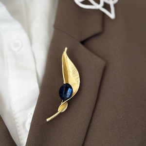 May include: A gold-coloured leaf-shaped brooch featuring a dark blue gemstone. The brooch is attached to a brown blazer, with a white collared shirt underneath. The design of the brooch is elegant and inspired by nature.