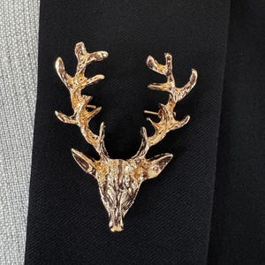May include: A gold-colored deer head brooch with detailed antlers. The brooch is pinned to a black fabric, showcasing its intricate design and metallic finish. This accessory adds a touch of nature-inspired elegance.