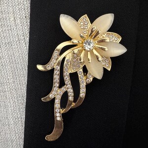May include: A gold-toned floral brooch with a central clear rhinestone and cream-colored petals. The brooch features a flowing, stylized design with additional clear rhinestones, set against a black background.
