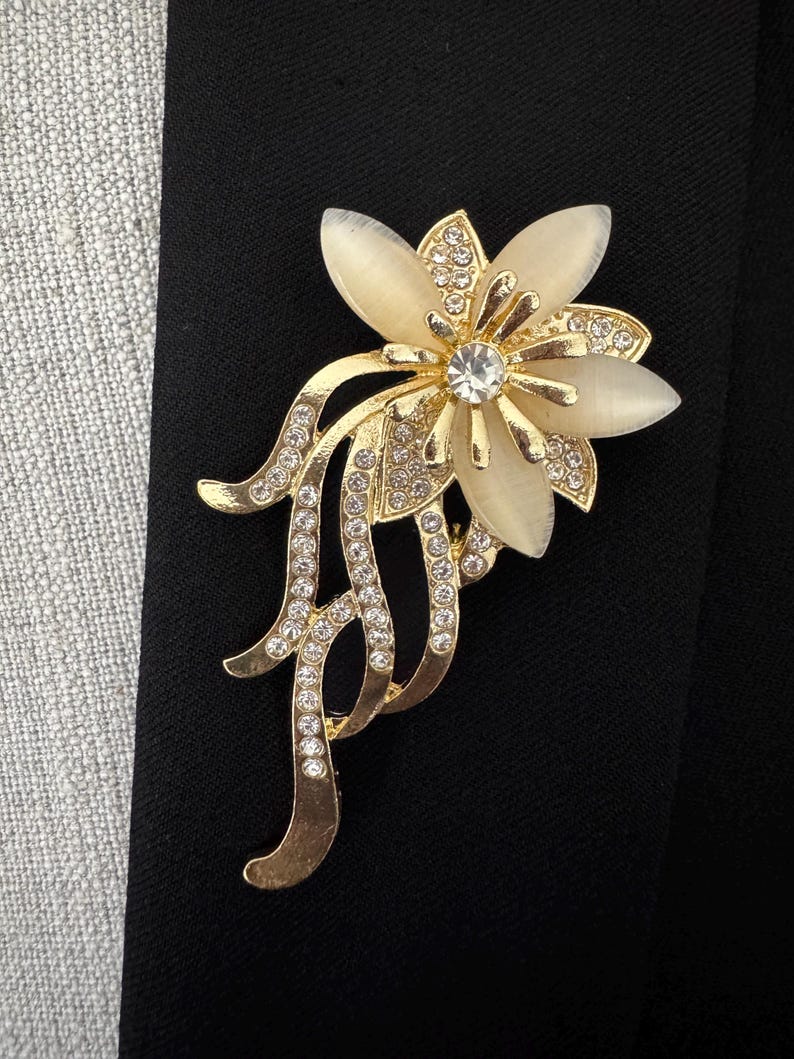 May include: A gold-toned floral brooch with a central clear rhinestone and cream-colored petals. The brooch features a flowing, stylized design with additional clear rhinestones, set against a black background.