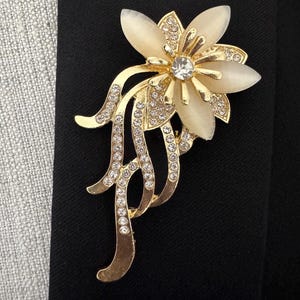 May include: A gold-toned floral brooch with a central clear rhinestone and cream-colored petals. The brooch features a detailed design with smaller rhinestones and flowing, decorative elements. The brooch is set against a black background.