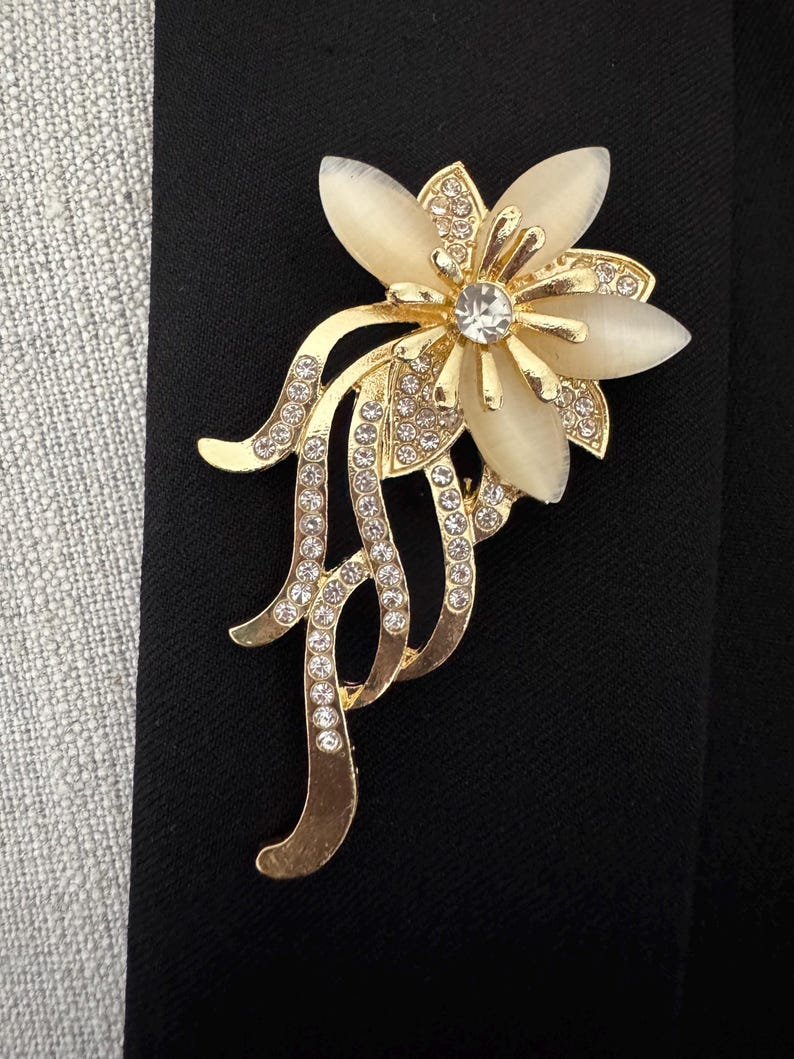 May include: A gold-toned floral brooch with a central clear rhinestone and cream-colored petals. The brooch features a detailed design with smaller rhinestones and flowing, decorative elements. The brooch is set against a black background.
