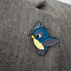Könnte beinhalten: Eine blaue und gelbe Vogelbrosche mit grauen Flügeln ist an einer grau-schwarzen Tweedjacke befestigt. Die Vogelbrosche hat ein cartoonartiges Design und besteht aus Emaille oder einem ähnlichen Material. Die Jacke hat einen klassischen, maßgeschneiderten Stil.