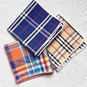 May include: Three folded handkerchiefs with plaid patterns. One is blue with red and white lines, another is tan with red and black lines, and the third is orange, blue, and red with a colorful plaid design. The handkerchiefs are on a gray surface.