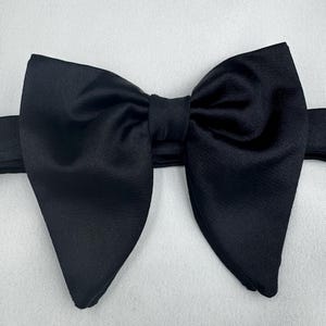 May include: A black bow tie with a classic butterfly shape. The bow tie is made of a smooth, solid black fabric and is set against a white background. The bow tie is a formal accessory.