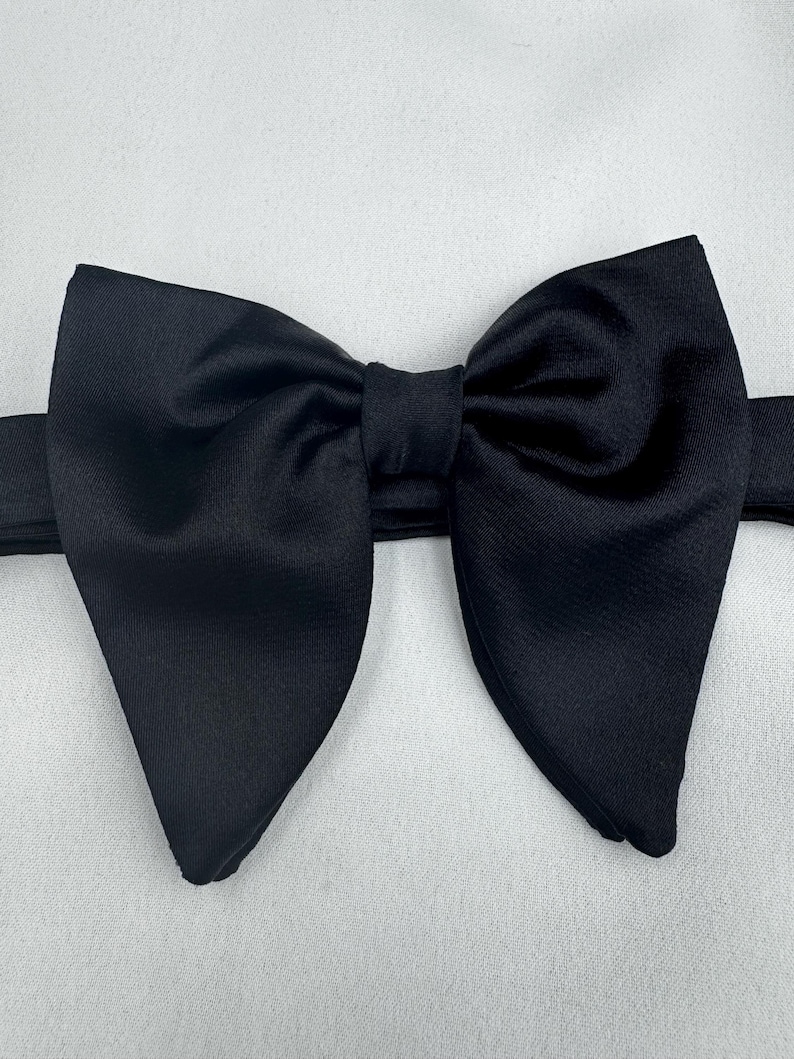 May include: A black bow tie in a traditional butterfly design. The bow tie is crafted from a smooth, solid black material and is displayed against a white backdrop. A formal accessory.