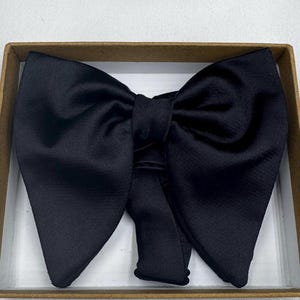 May include: A black bow tie made from a glossy material, displayed in a brown cardboard box. The bow tie is well-shaped and designed for formal occasions. The fabric has a subtle sheen.