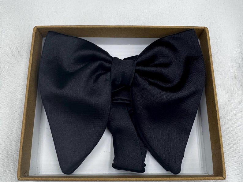 May include: A black bow tie made from a glossy material, displayed in a brown cardboard box. The bow tie is well-shaped and designed for formal occasions. The fabric has a subtle sheen.
