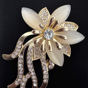 May include: A gold-toned flower brooch with a central clear crystal and cream-colored petals. The brooch features multiple clear crystals and curved gold accents, set against a black background.